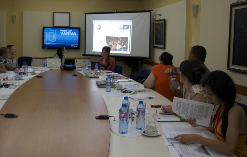 a group of people sit around a conference table as the man at the screen speaks on a computer screen is on