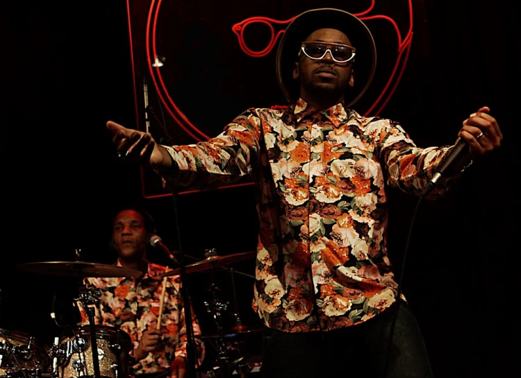 a man wearing sunglasses and flower shirt standing in front of a drum kit