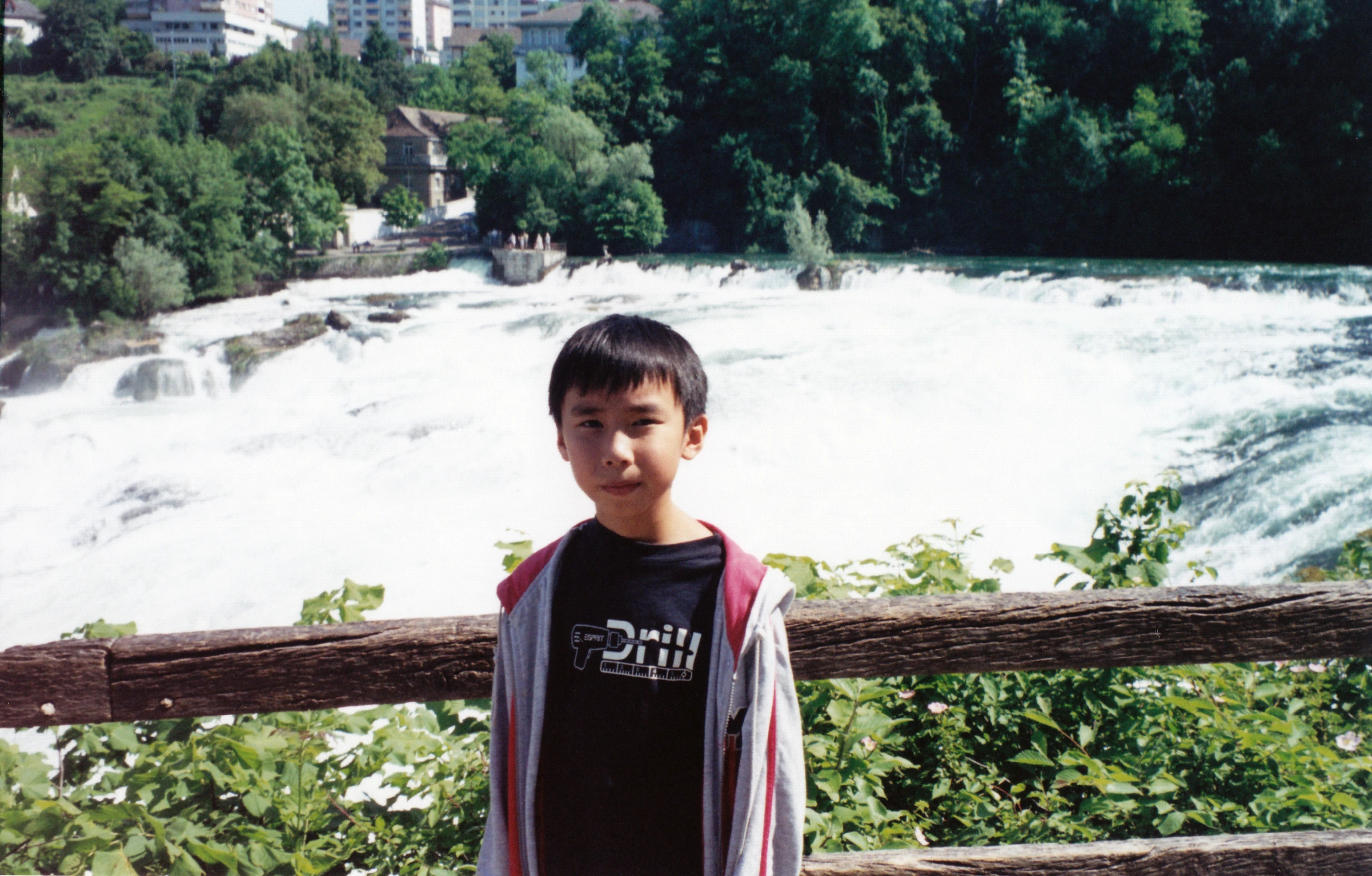 a boy in a sweatshirt standing on a wooden fence looking over a river