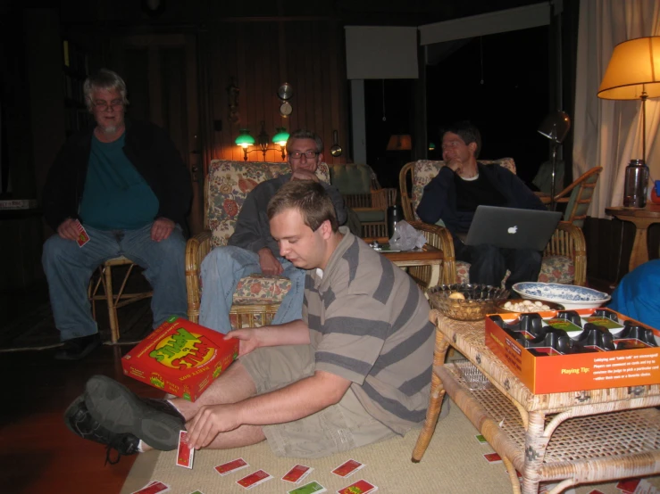 there are men sitting in the living room, a man is opening a box with cards