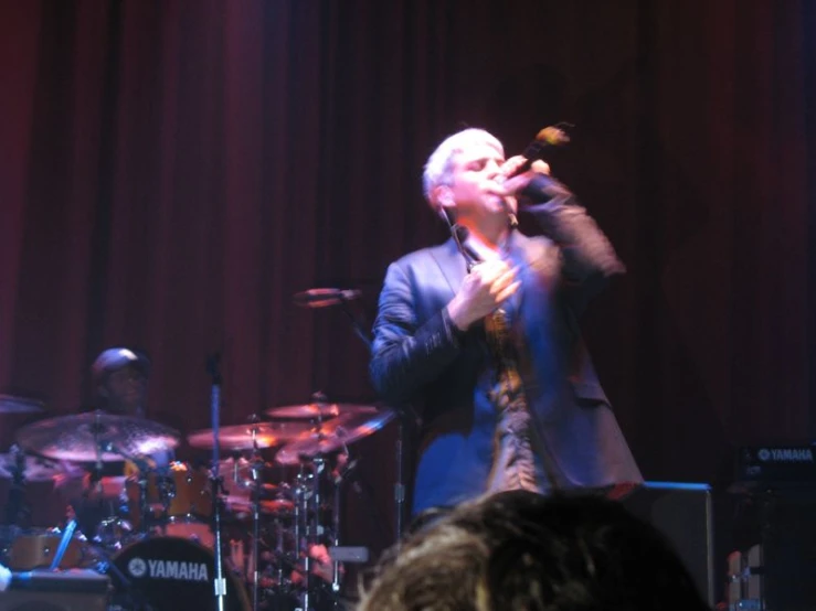 a man with white hair sings in front of several drummers