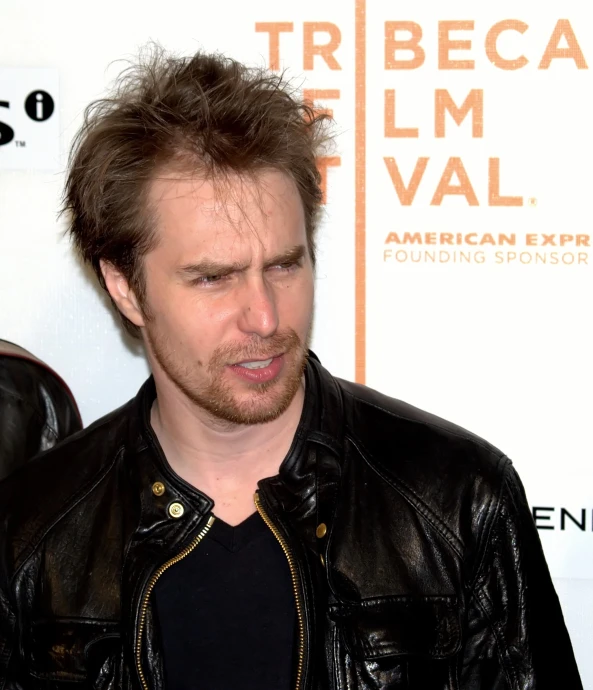 a man with short brown hair and a black shirt standing in front of a banner