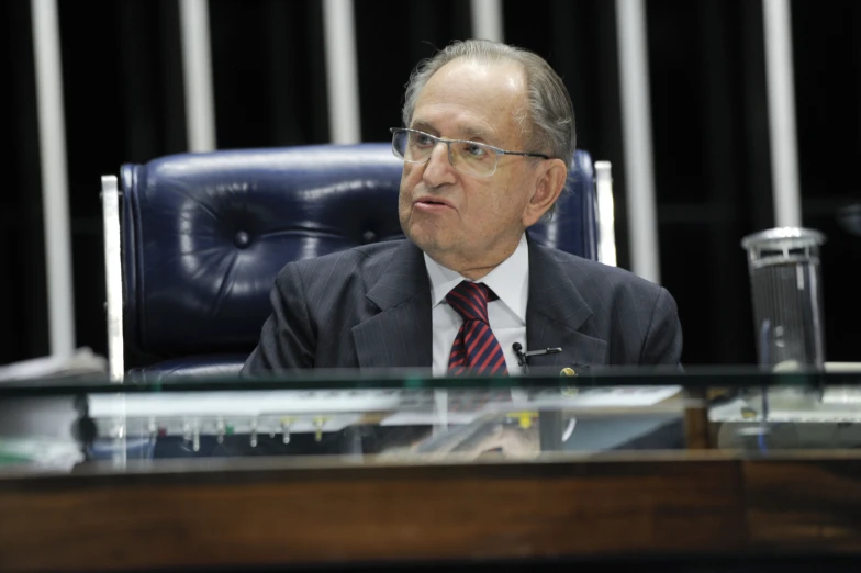 a man wearing glasses, a suit and tie is sitting down at a desk