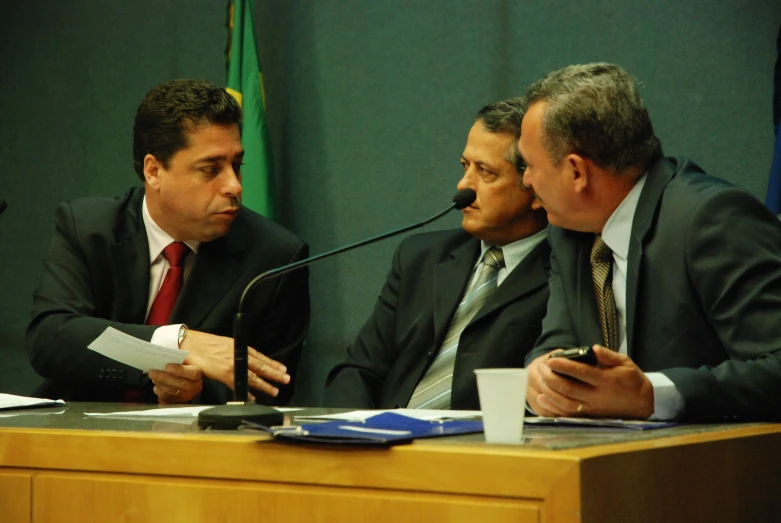 three men in suits sit around a table while looking at papers