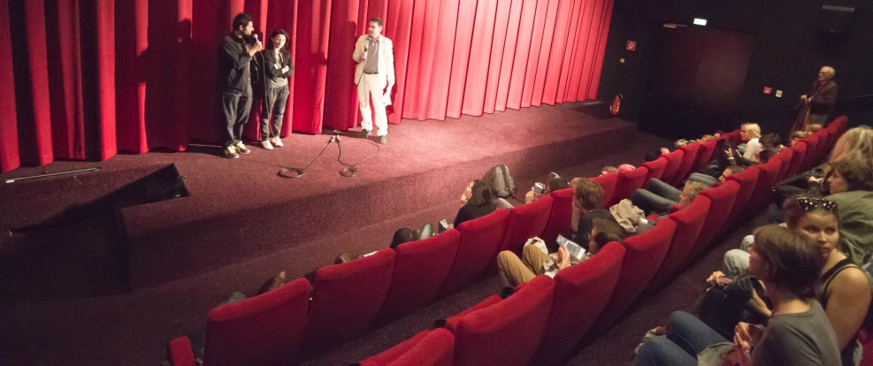 people standing and sitting on red chairs in a theater