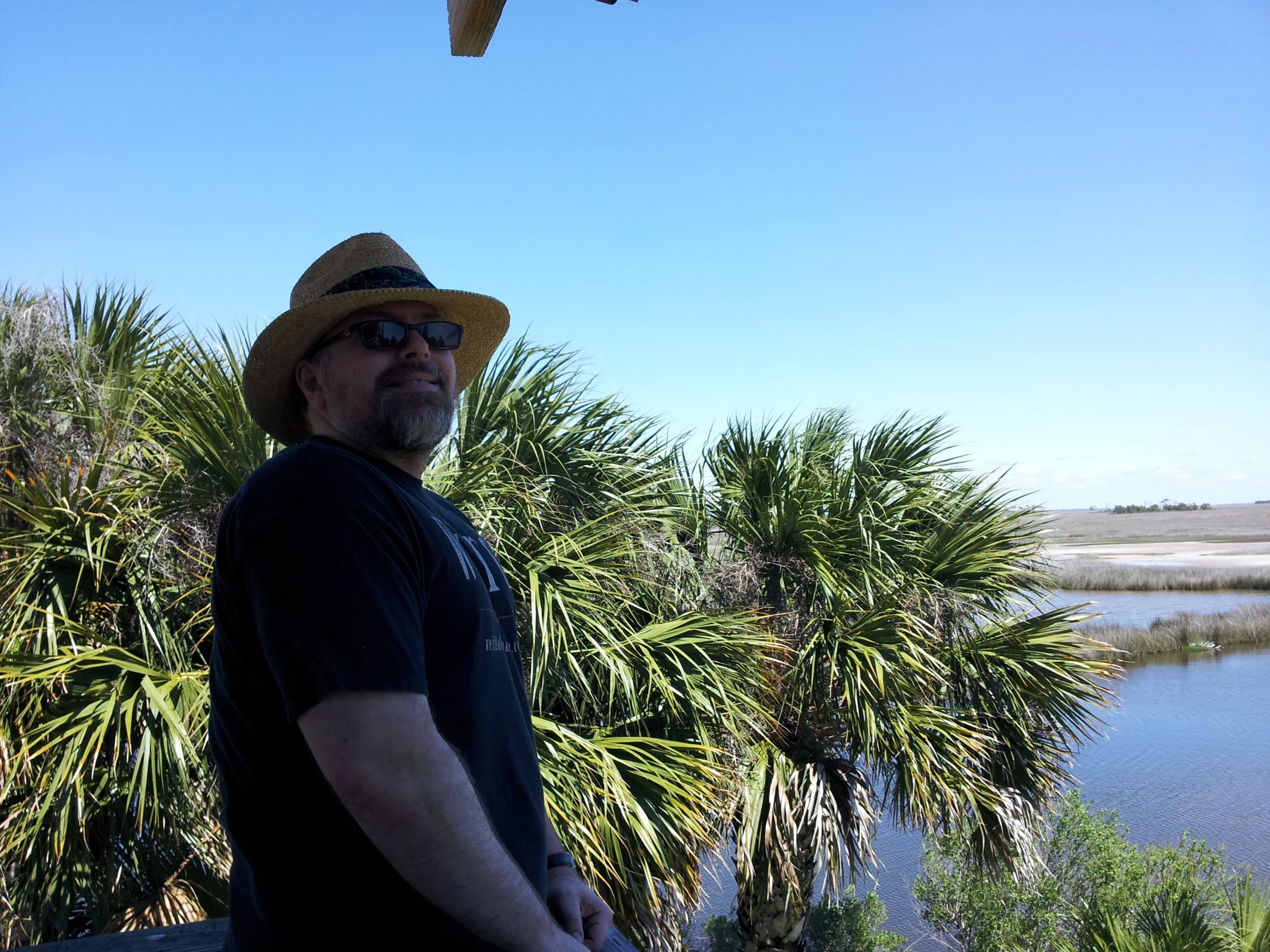 a man wearing a hat stands on the balcony in front of a lake and palm trees