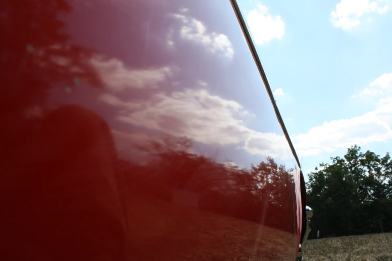 trees against blue sky and clouds reflected in a red body of car