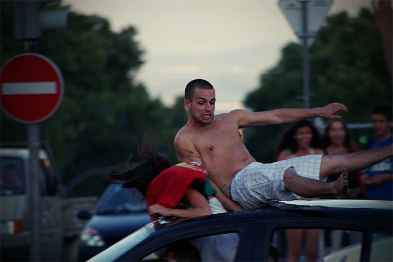 a young man with  is dancing on top of a car in the street