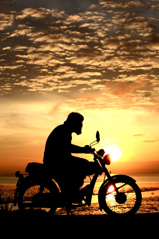 a silhouetted person riding a motorcycle on a beach with the sun setting