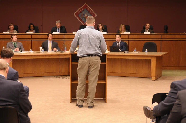 a man stands before two wooden tables with people seated