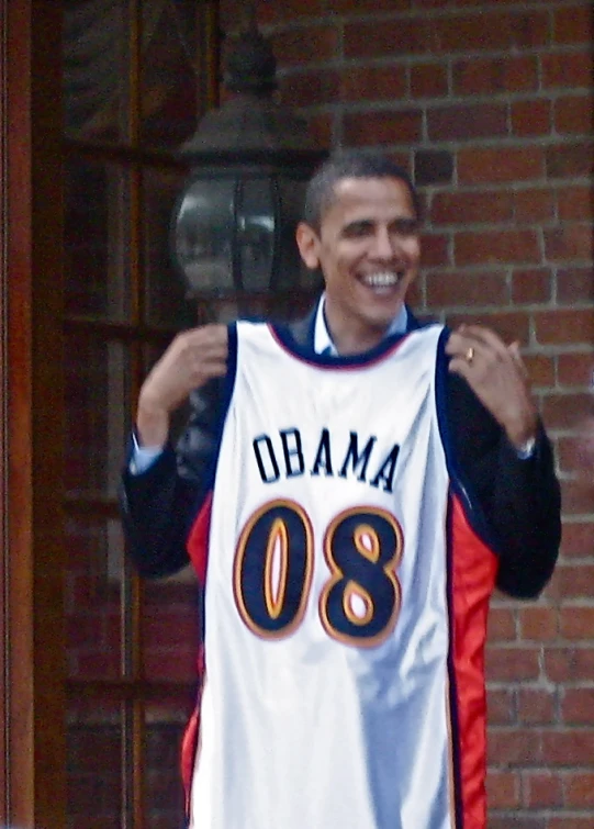 barack obama holding up a basketball uniform in front of a brick building