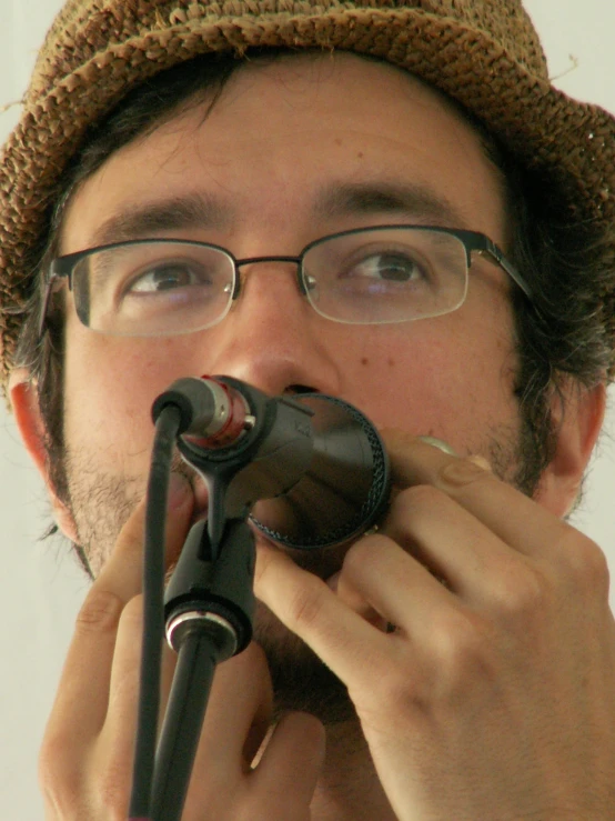 a man wearing glasses holds his mouth to the side while standing with his hat and microphone in front of his mouth