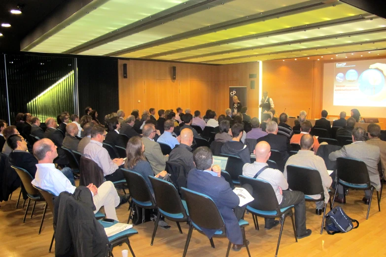 a large audience of people sitting at tables in front of a presentation screen