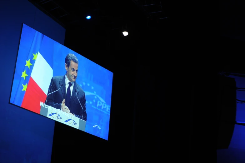 a man standing at a lectern in front of a large screen