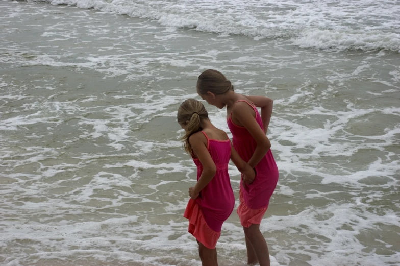 two girls in pink dresses stand in the surf at the beach