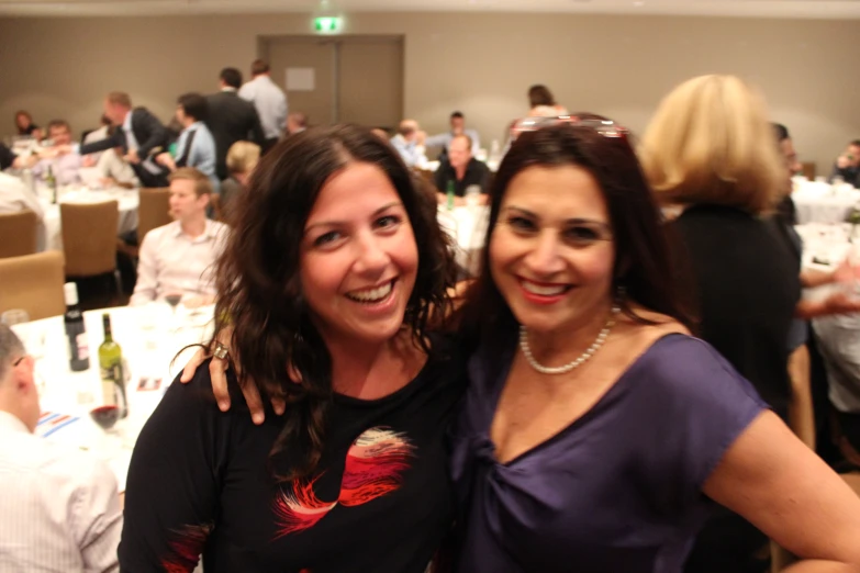 two women smile for the camera as they sit at tables with other people