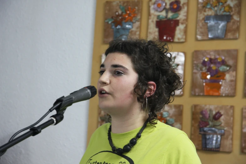 woman in yellow shirt singing into microphone near wall with glass artwork on it