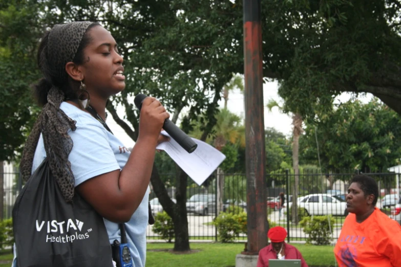 a black woman holding a microphone and papers with other people watching