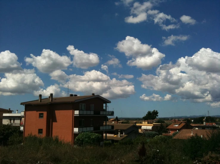 a small brown house surrounded by greenery in front of a blue sky with white clouds
