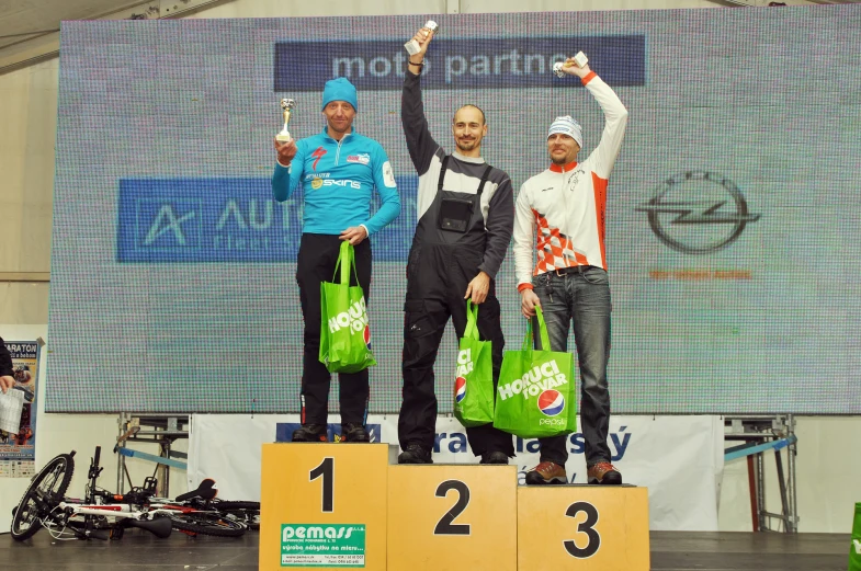 three men standing on two winners pedestals holding up green bags