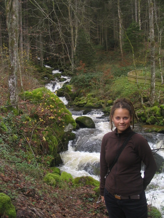 a woman in a brown shirt posing on a moss covered bank near a waterfall