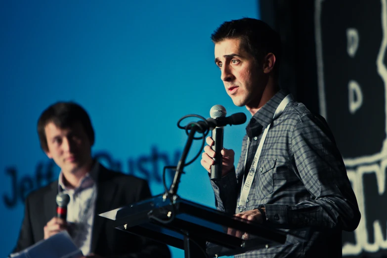 two men standing behind a podium, one speaking and the other standing behind a microphone