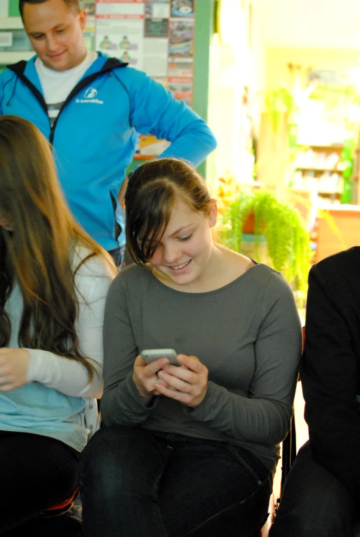 a girl sitting in a chair looking at her phone with other people standing around