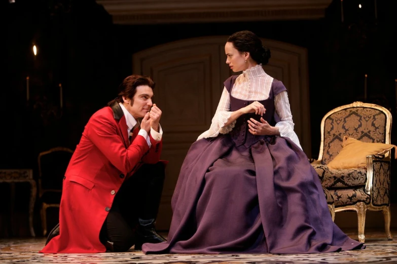 two actors dressed in period costumes sit on stage