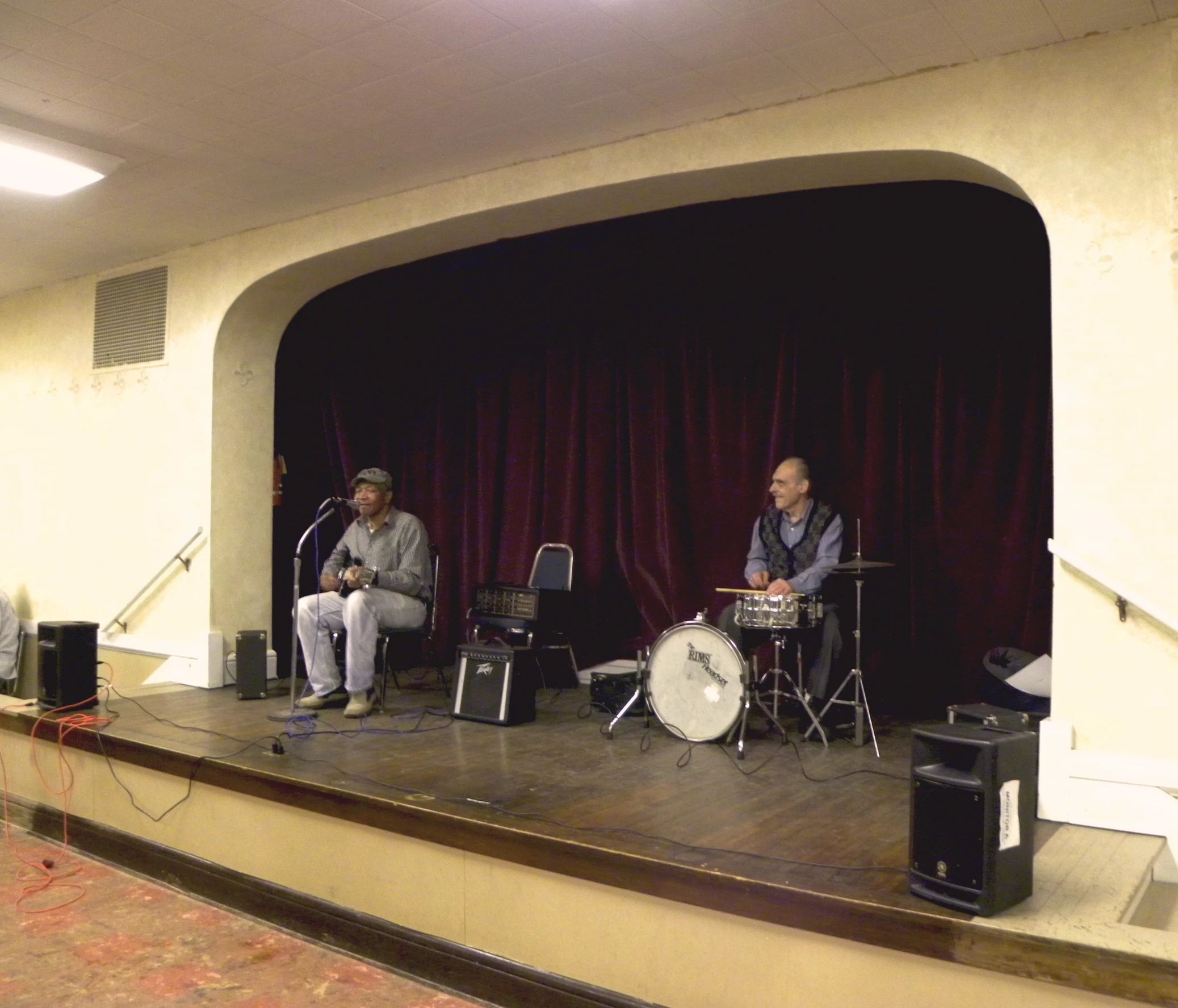 two men in blue shirts sit on stage while one man plays a drumset