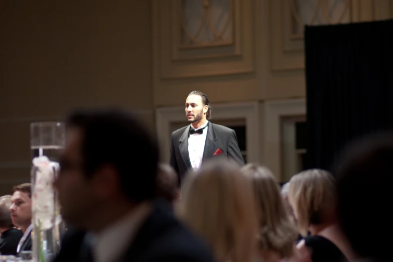 man wearing black tuxedo in middle of audience at formal dinner