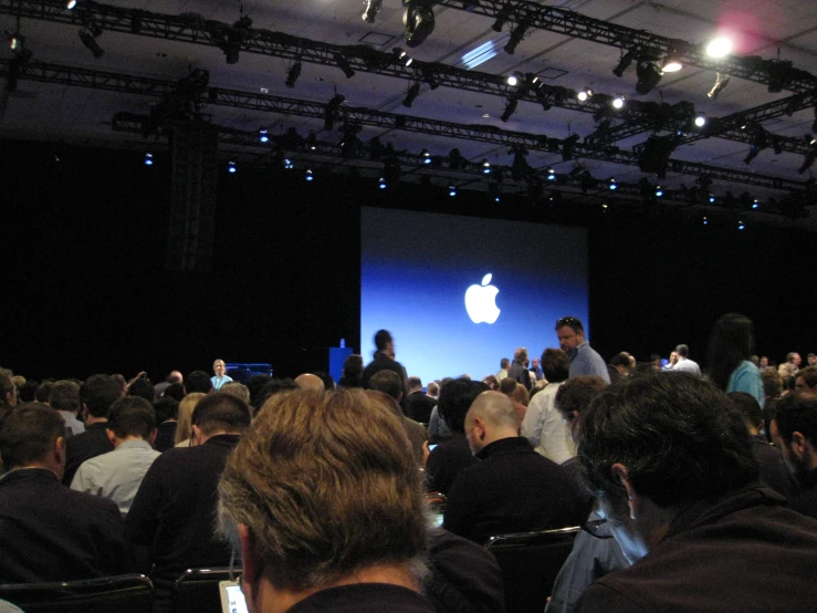 a crowd in a building standing around and looking at the stage with an apple logo projected