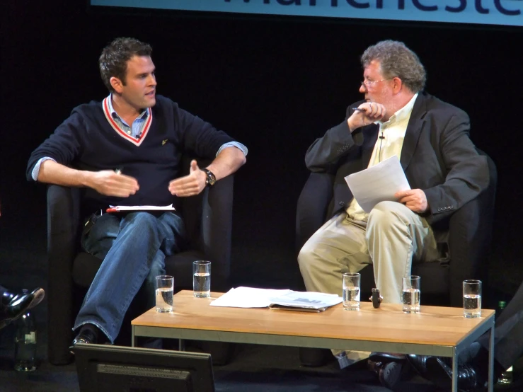 two men sitting down talking on chairs in front of a stage