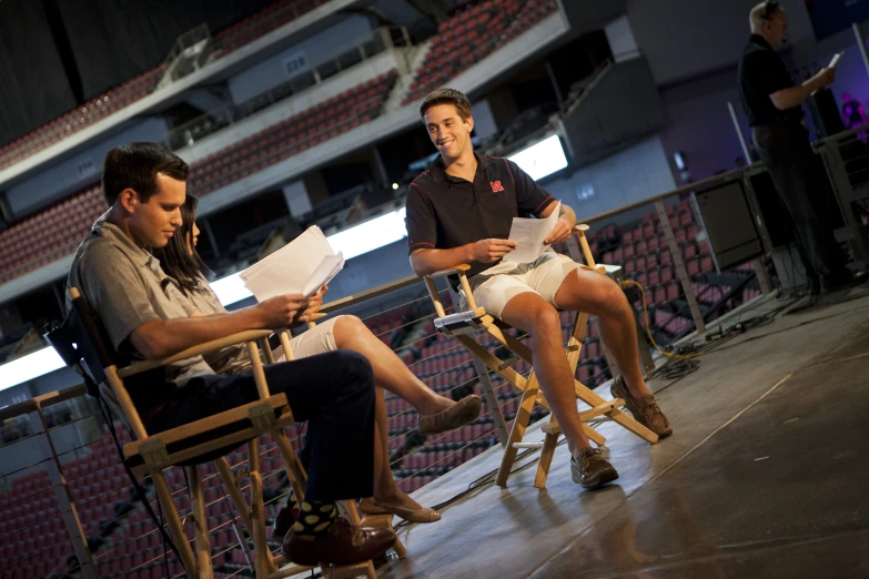 two men sitting on wooden folding chairs in front of an audience