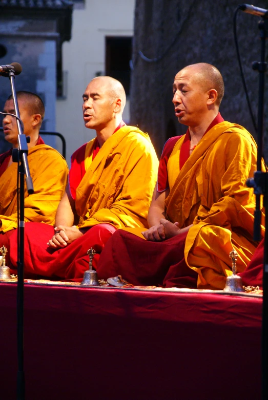 three monks in yellow robes sit on stage together