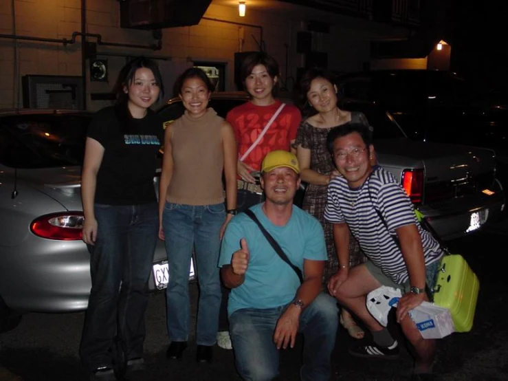 four people are standing in front of a parked car and one woman has her thumbs up