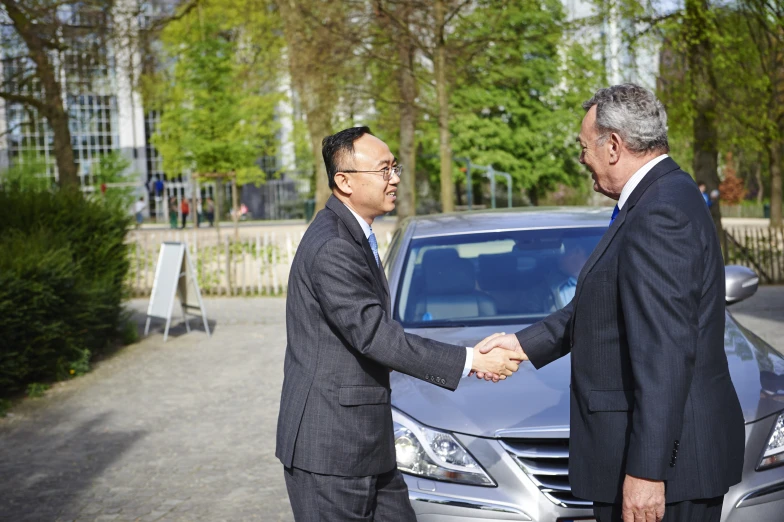 two men in suits shaking hands next to a car