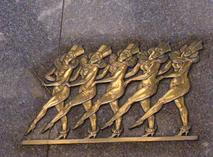 bronze artwork on the side of a building with many ladies in gold outfits holding onto two spears