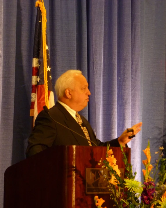 a man in a black suit sitting at a podium with flowers