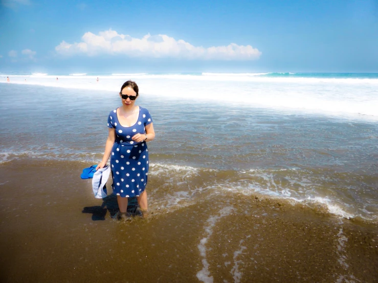 a woman with sunglasses on, standing in shallow water on the beach