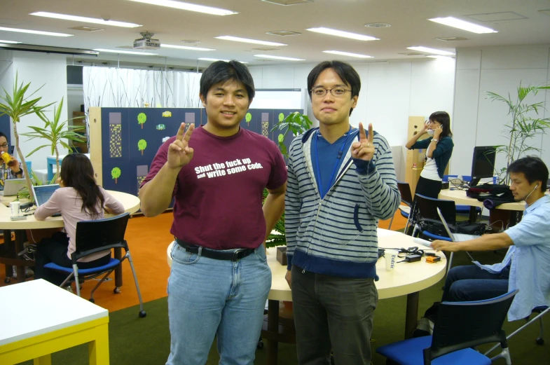 two young men standing in an office room holding up their thumbnails