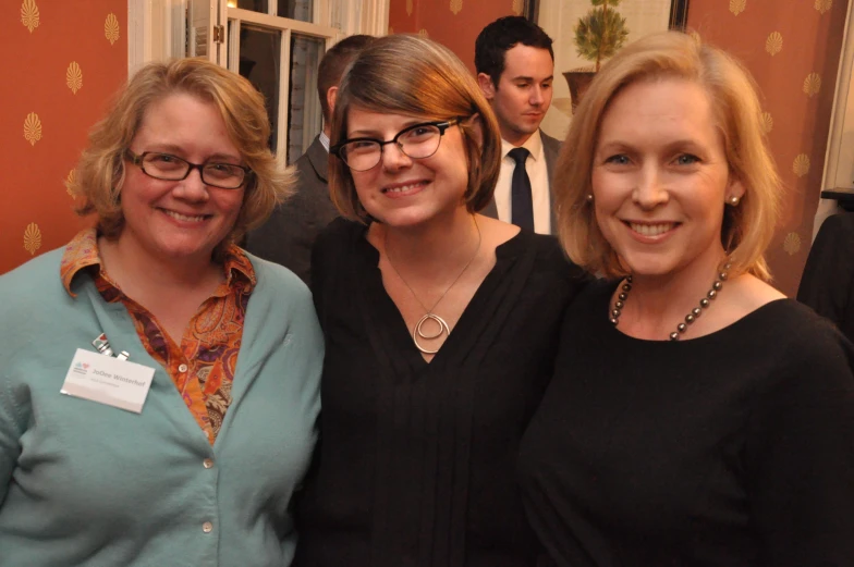 three women smiling in a room with other men and women in the background