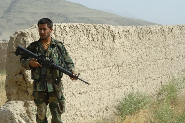 a soldier holding a rifle next to a rock wall