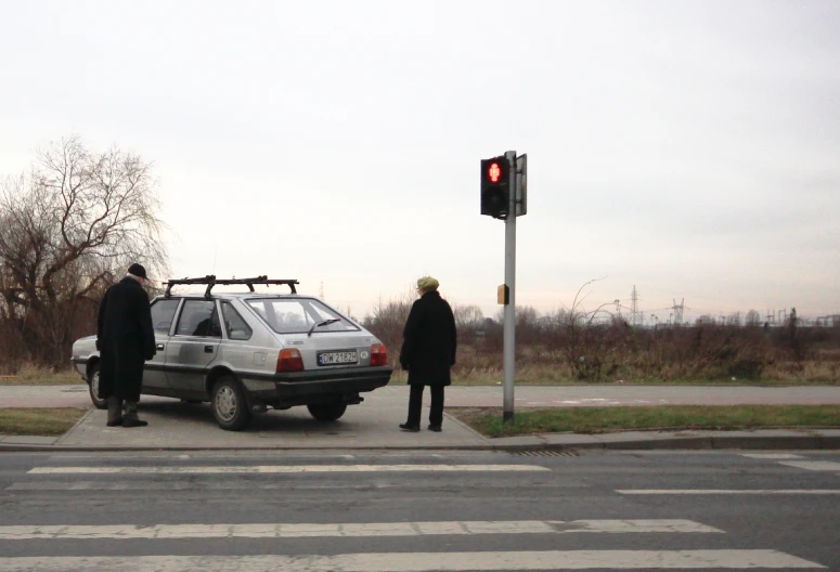 two men stand by a car at an intersection, watching another man drive