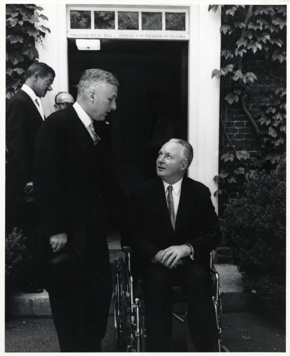 two men in suits stand outside an entrance with one man in a wheel chair and another man in a suit behind him