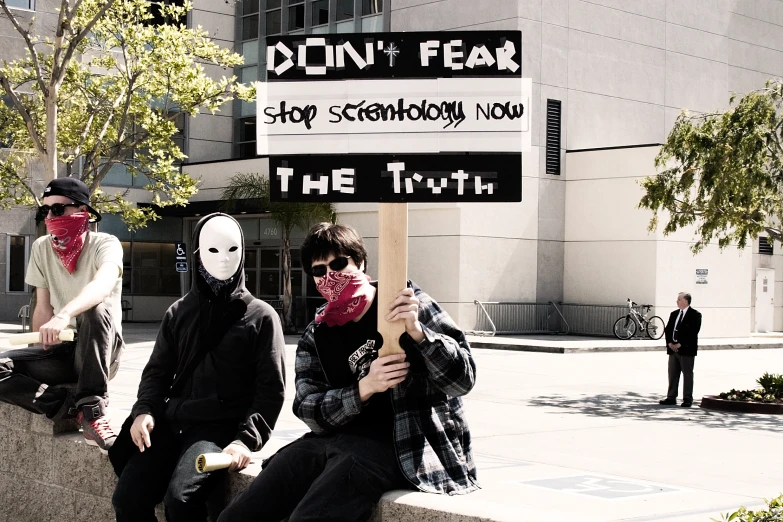 three people wearing face coverings sit on the curb of a street