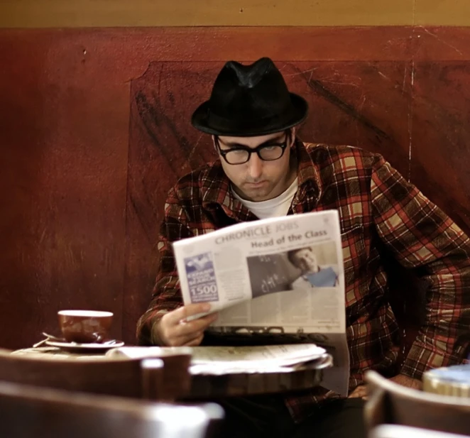 a man wearing glasses and a hat sits at a table with a newspaper