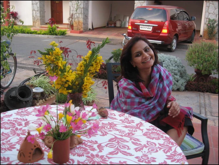 a woman is smiling while sitting at an outside table with flowers in a vase