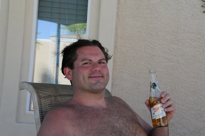 a shirtless man holding up a bottle and sitting on a chair