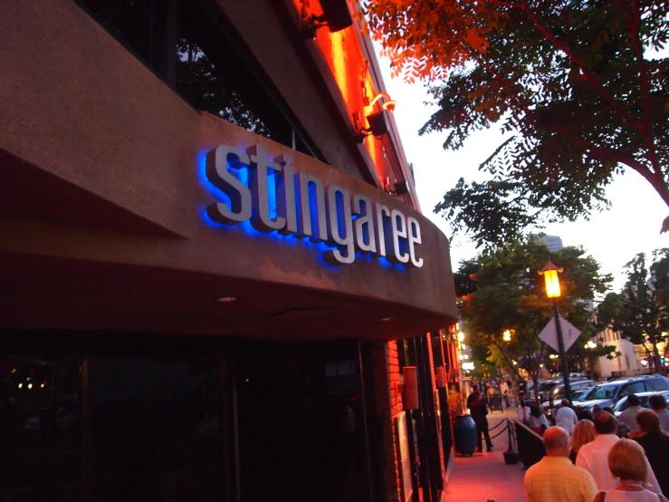 people walk down the sidewalk below the large blue lighted sign on the building