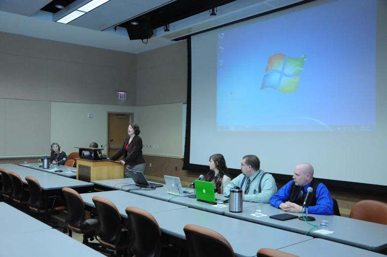 people sitting at tables in front of a screen, and some are using laptops